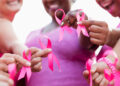 Cropped view of a multi-ethnic group of women of mixed ages standing together outdoors, wearing pink, at a breast cancer awareness rally, raising money to find a cure. They are each holding a breast cancer awareness ribbon. The focus is on their hands.