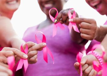 Cropped view of a multi-ethnic group of women of mixed ages standing together outdoors, wearing pink, at a breast cancer awareness rally, raising money to find a cure. They are each holding a breast cancer awareness ribbon. The focus is on their hands.